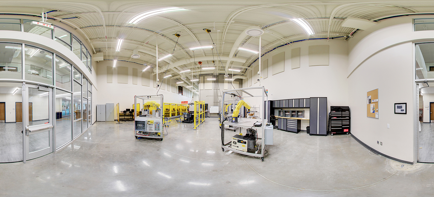 Panoramic view of an advanced manufacturing lab with high ceilings, featuring industrial robotic arms, yellow safety enclosures, control stations, and tool cabinets arranged across a spacious, well-lit training environment.