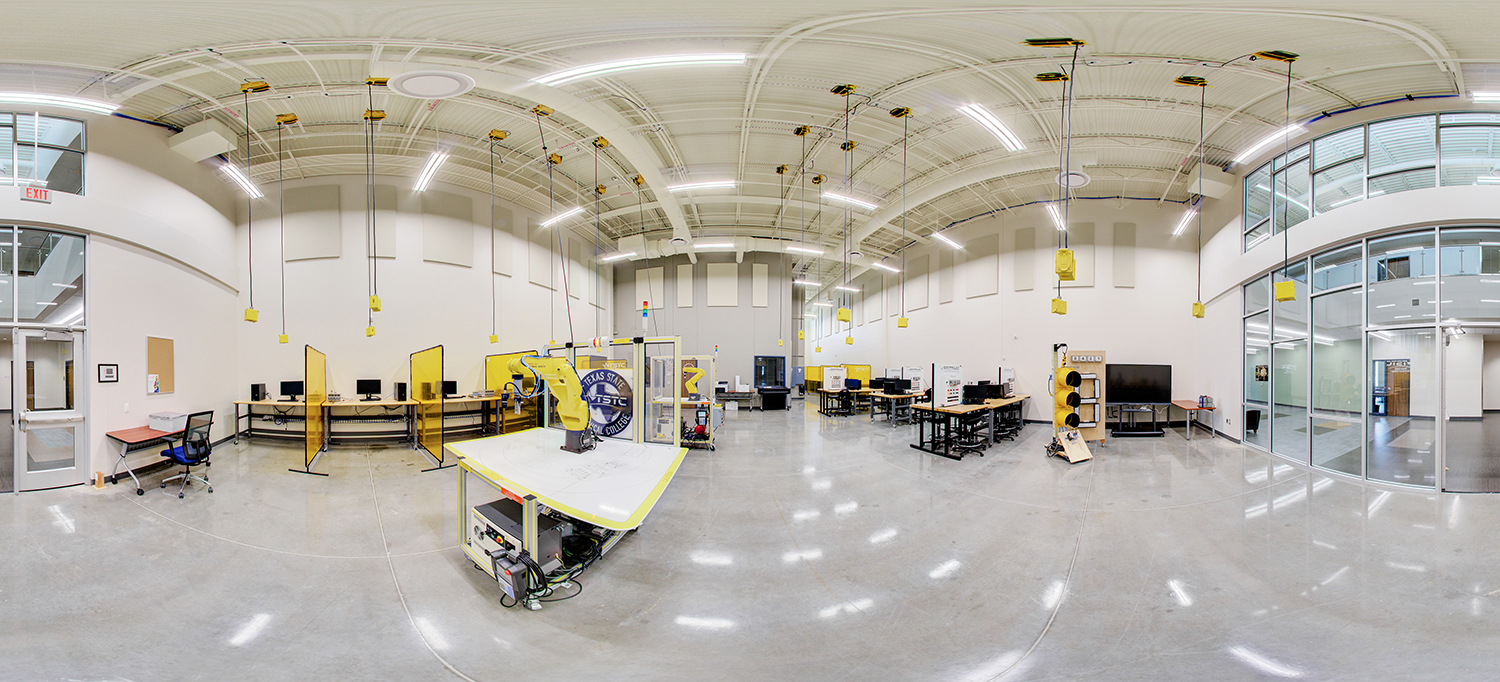 Panoramic view of a robotics and automation training lab with high ceilings, featuring robotic arms inside yellow safety enclosures, computer workstations along the walls, suspended power drops from the ceiling, and an open, well-lit workspace.