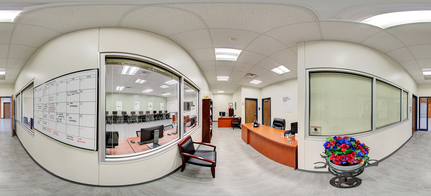 Panoramic view of an office and computer lab area, featuring a reception desk, a wall-mounted calendar whiteboard, and adjacent rooms visible through interior windows, including a classroom with rows of desktop computers.