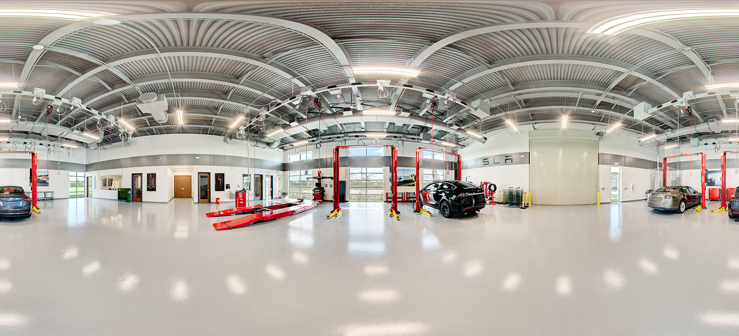 Panoramic view of a modern automotive training lab featuring multiple vehicle lifts, red service platforms, and several cars positioned for maintenance, with high ceilings, bright lighting, and organized tool and equipment areas throughout the spacious facility.