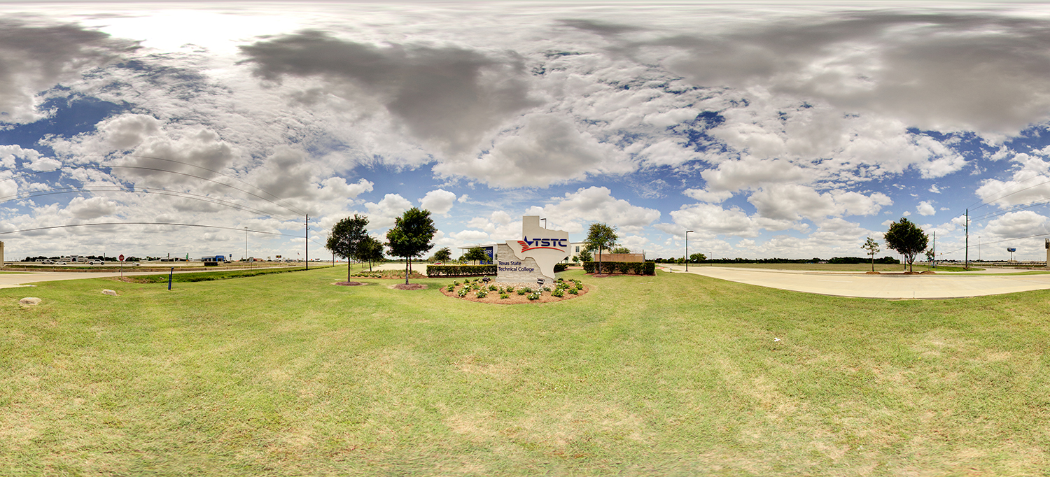 Panoramic view of a Texas State Technical College entrance sign on a landscaped grassy area with small trees and surrounding roads, set under a wide sky filled with large, scattered clouds.