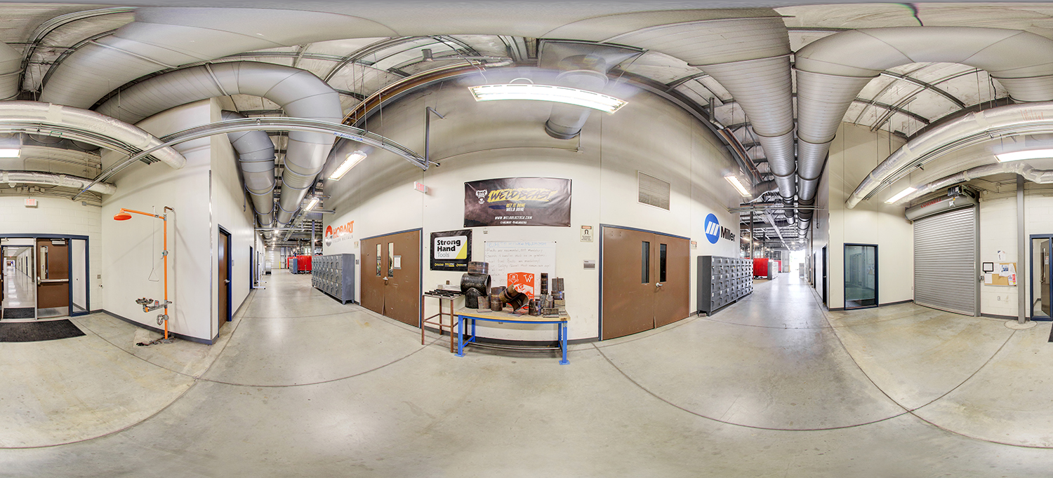 Panoramic view of a welding training facility corridor with high ceilings and exposed ventilation ducts, featuring rows of lockers, workshop doors, safety showers, and equipment displays, with welding booths visible further down the hall.