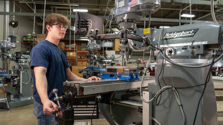 A student wearing safety glasses stands at a Bridgeport milling machine, adjusting the controls while a metal piece is secured in a vise on the worktable in a machine shop.