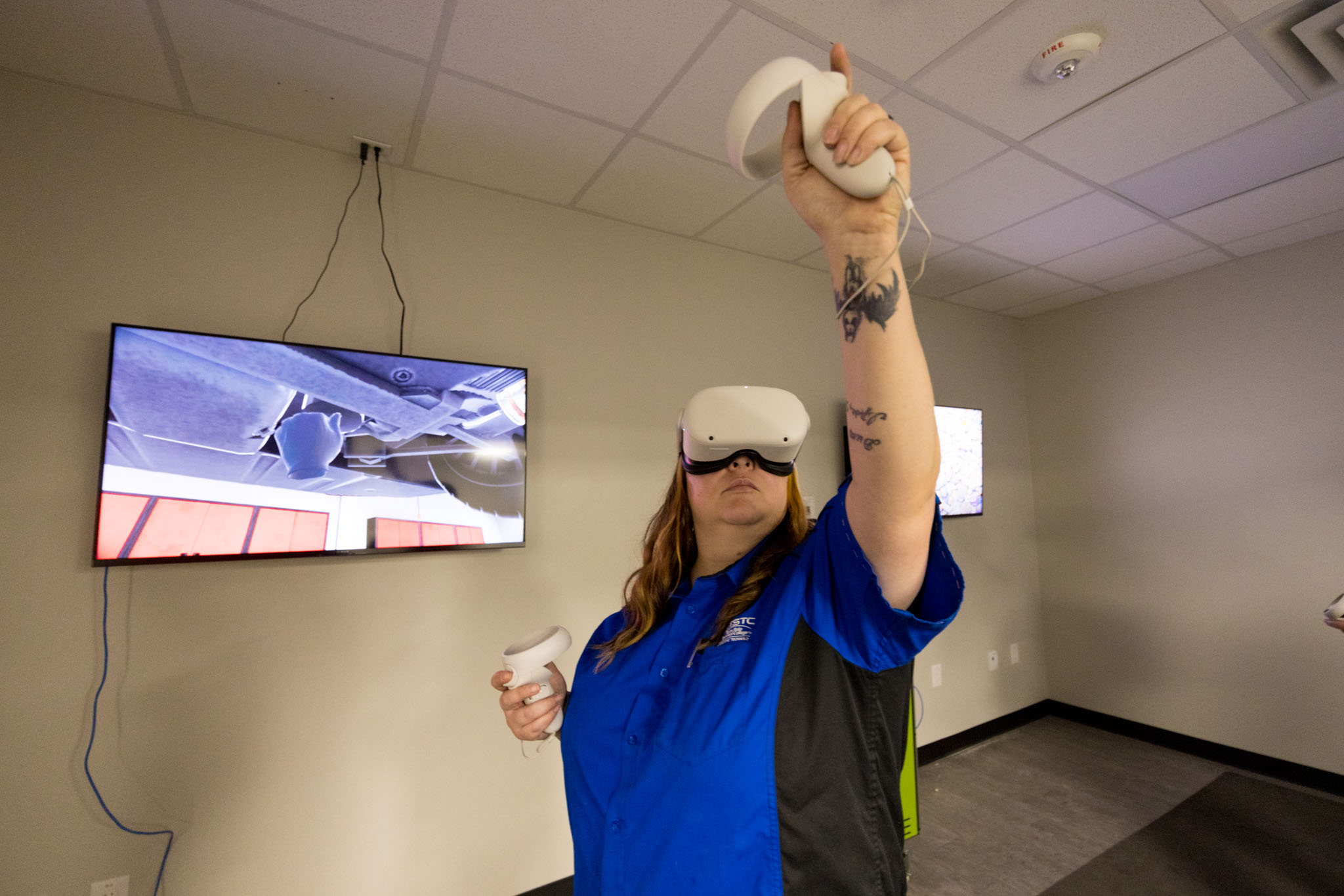 A person wearing a virtual reality headset holds controllers and raises one hand while interacting with a VR simulation, with a display screen showing the virtual environment behind them.