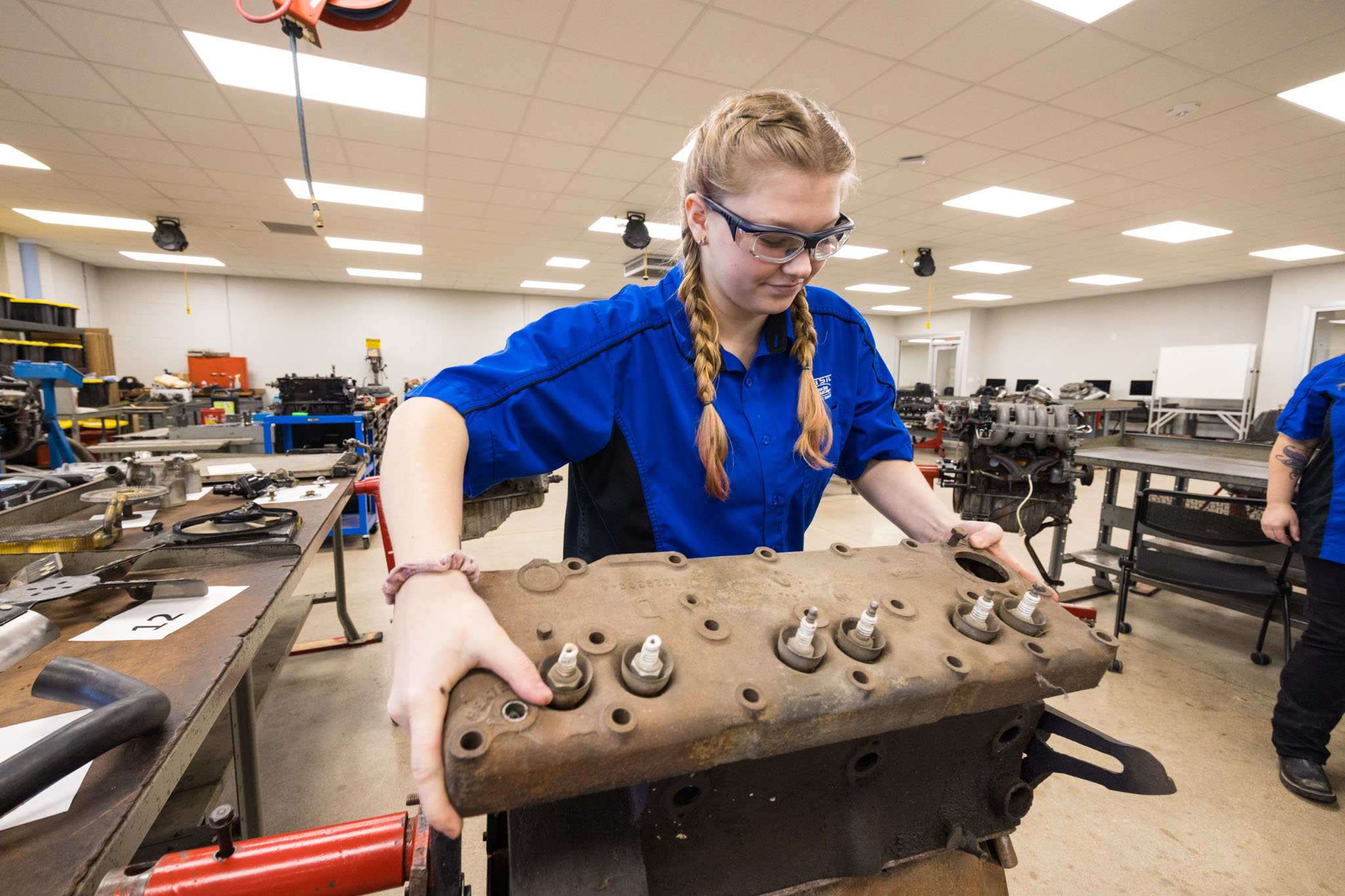 A student wearing safety glasses and a blue work shirt lifts and positions a heavy engine cylinder head with visible spark plugs in an automotive lab.
