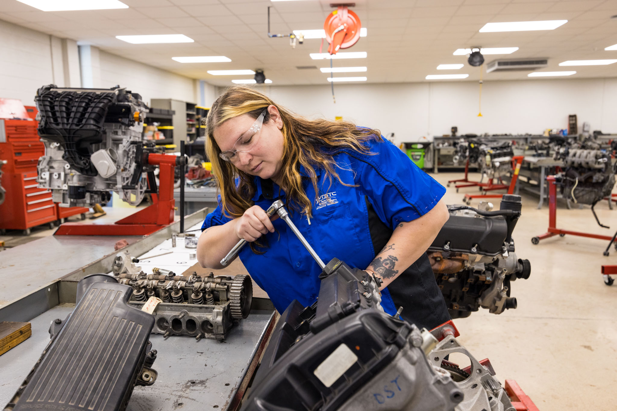 A student wearing safety glasses and a blue work shirt uses a ratchet to tighten a component on an engine assembly, working at a bench in an automotive lab surrounded by engine parts.