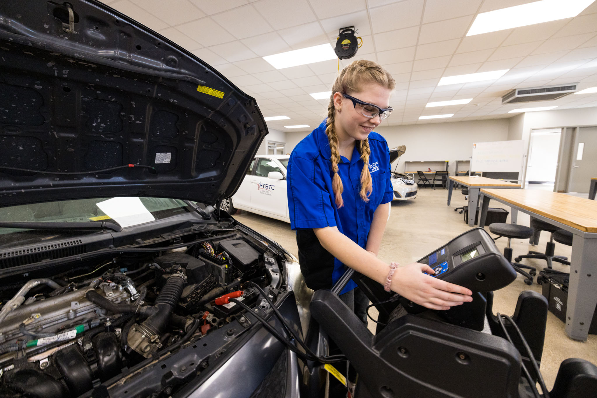 A student wearing safety glasses uses a diagnostic machine connected to a car with its hood open, checking vehicle systems in an automotive lab.