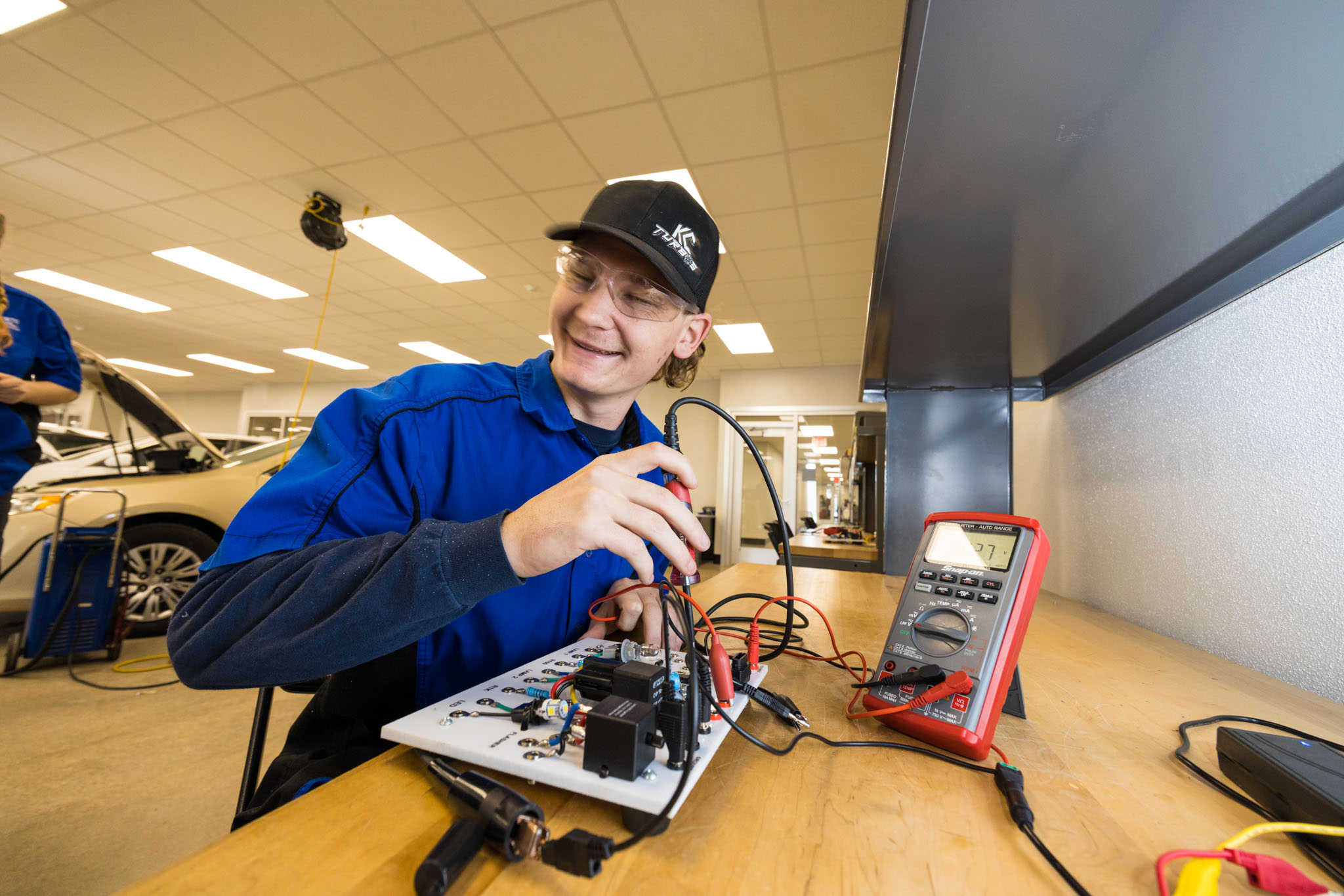 A student wearing safety glasses and a cap uses a multimeter to test an electrical circuit board on a workbench, smiling while taking measurements in a lab.