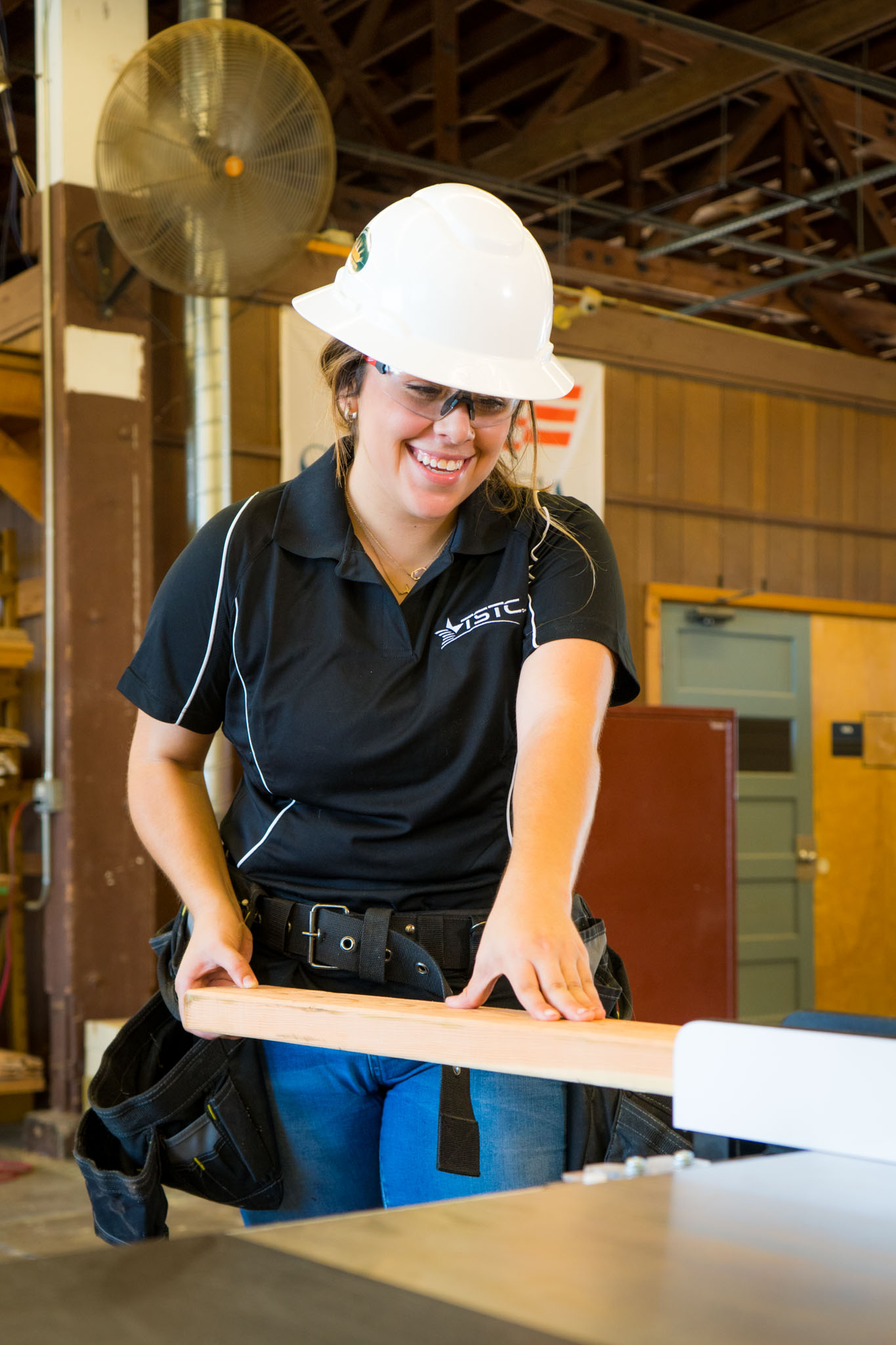 In a workshop, a student wearing a white hard hat and safety glasses holds and aligns a wooden board on a table saw, preparing it for a precise cut.