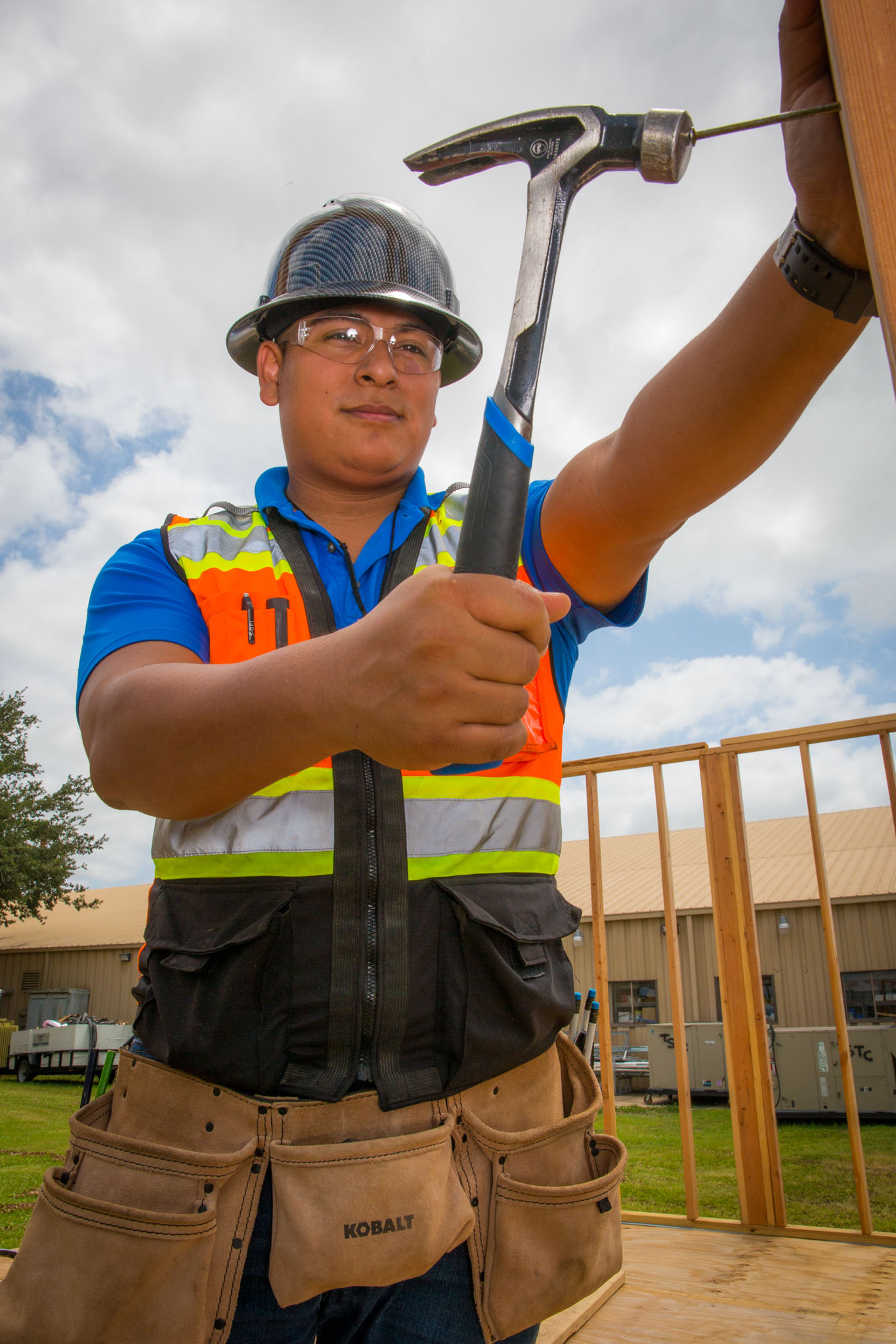 At an outdoor construction site, a student wearing a hard hat, safety glasses, and a reflective vest uses a hammer to drive a nail into a wooden frame, with partially built walls behind him.