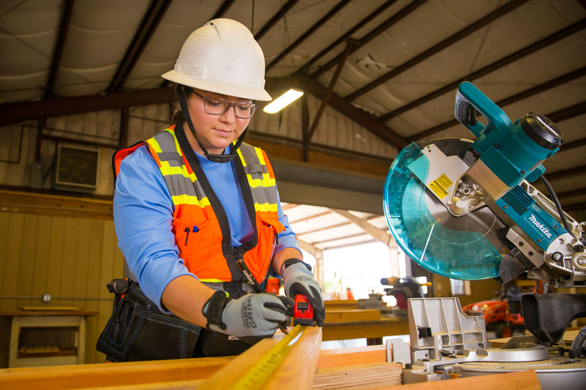 In a workshop, a student wearing a hard hat, safety glasses, and a reflective vest measures a wooden board with a tape measure beside a miter saw, preparing it for a precise cut.