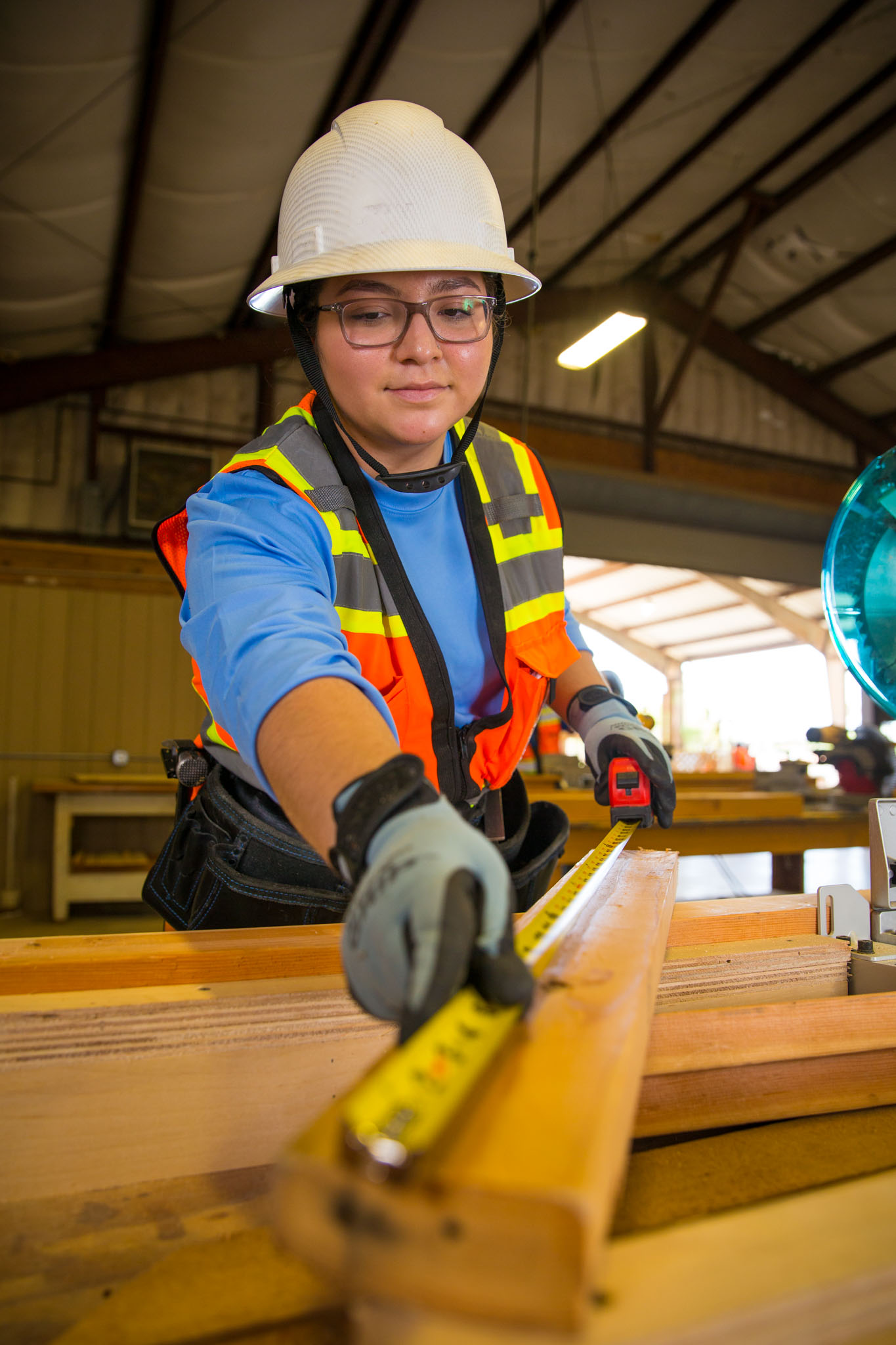 In a workshop, a student wearing a hard hat, safety glasses, and a reflective vest measures a wooden board with a tape measure, focusing closely on the marked length before cutting.