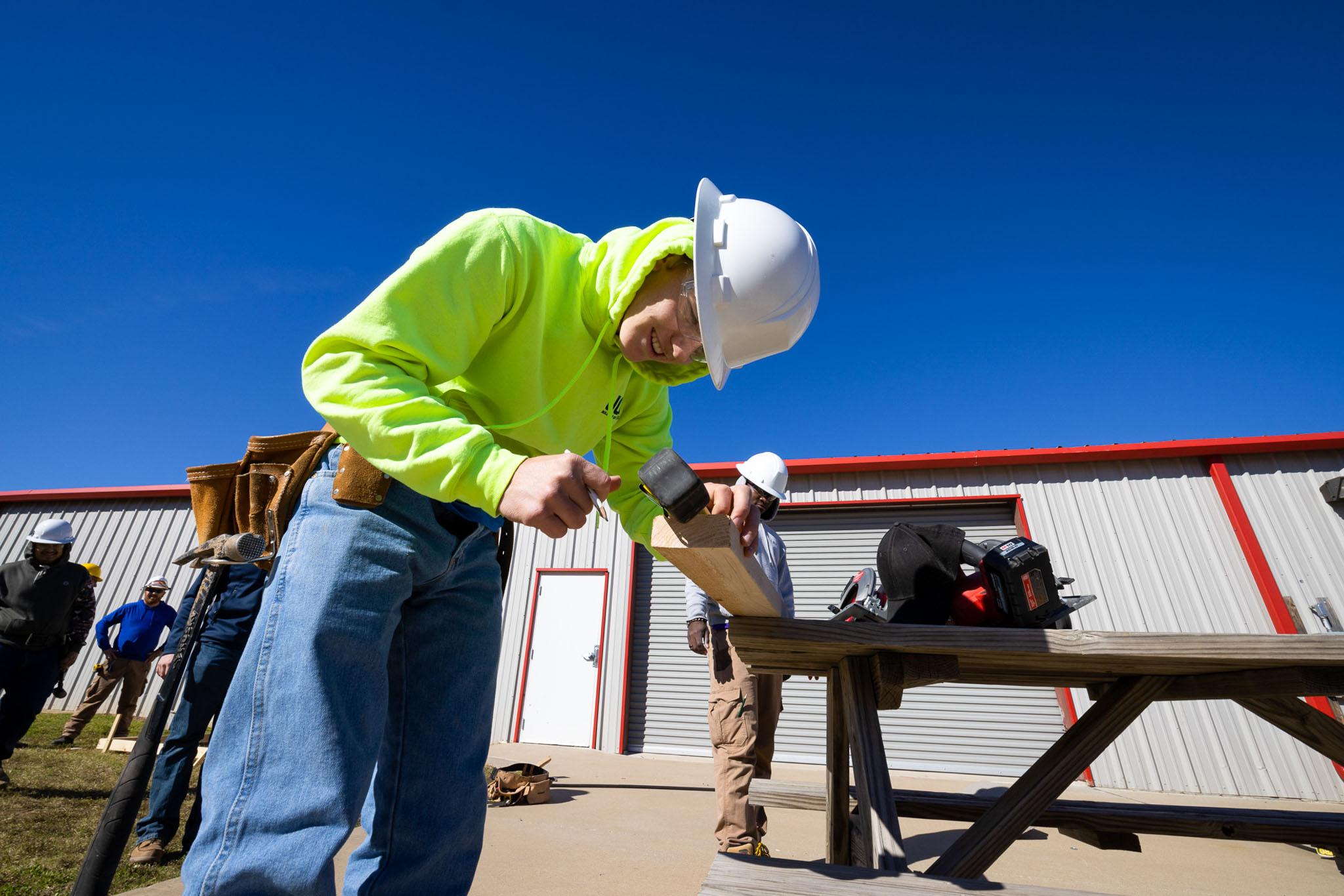 At an outdoor training site, a student wearing a white hard hat and neon safety hoodie uses a hammer to drive a nail into a wooden board on a workbench, with other students and a workshop building in the background.