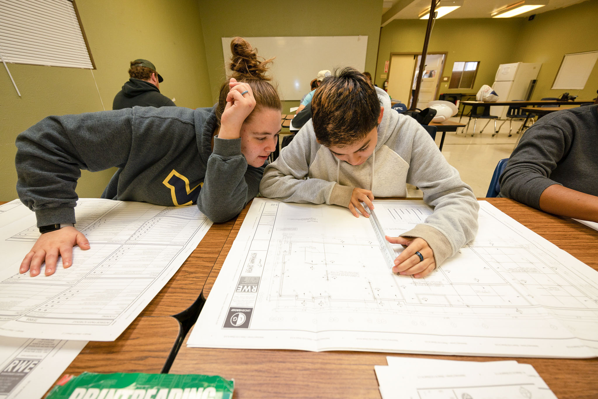 In a classroom, two students review a large set of technical drawings, with one using a ruler to measure and trace lines while the other leans in to discuss the plans.