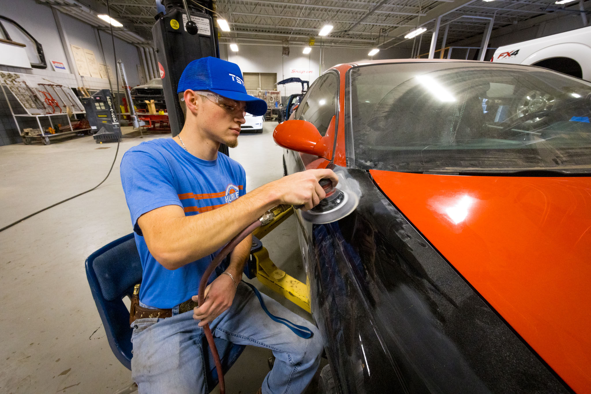 Student uses a powered sander to smooth the surface of a car door, removing paint on a red and black vehicle inside an auto body workshop.