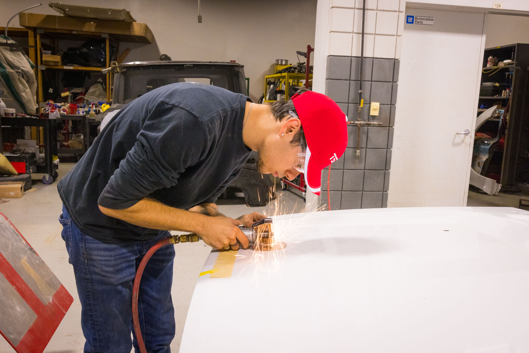 Student wearing a red cap and safety glasses uses a handheld grinder on a car panel, producing sparks while smoothing the metal surface in an auto shop.