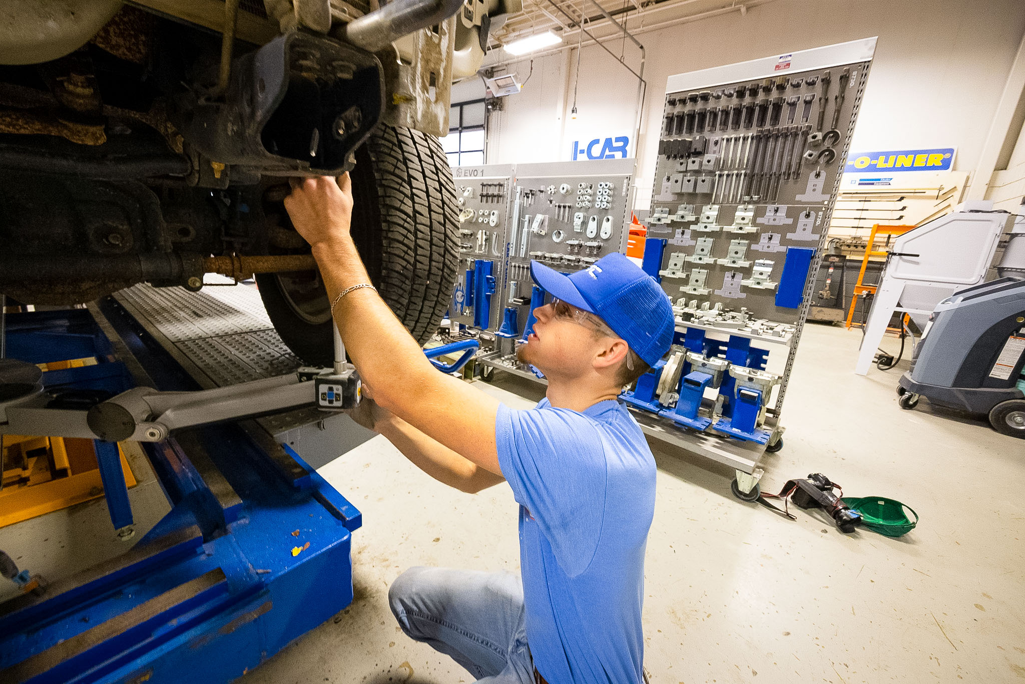 Student wearing a blue cap inspects the underside of a raised vehicle, reaching toward the suspension near a tire in an auto repair training shop with organized tools in the background.