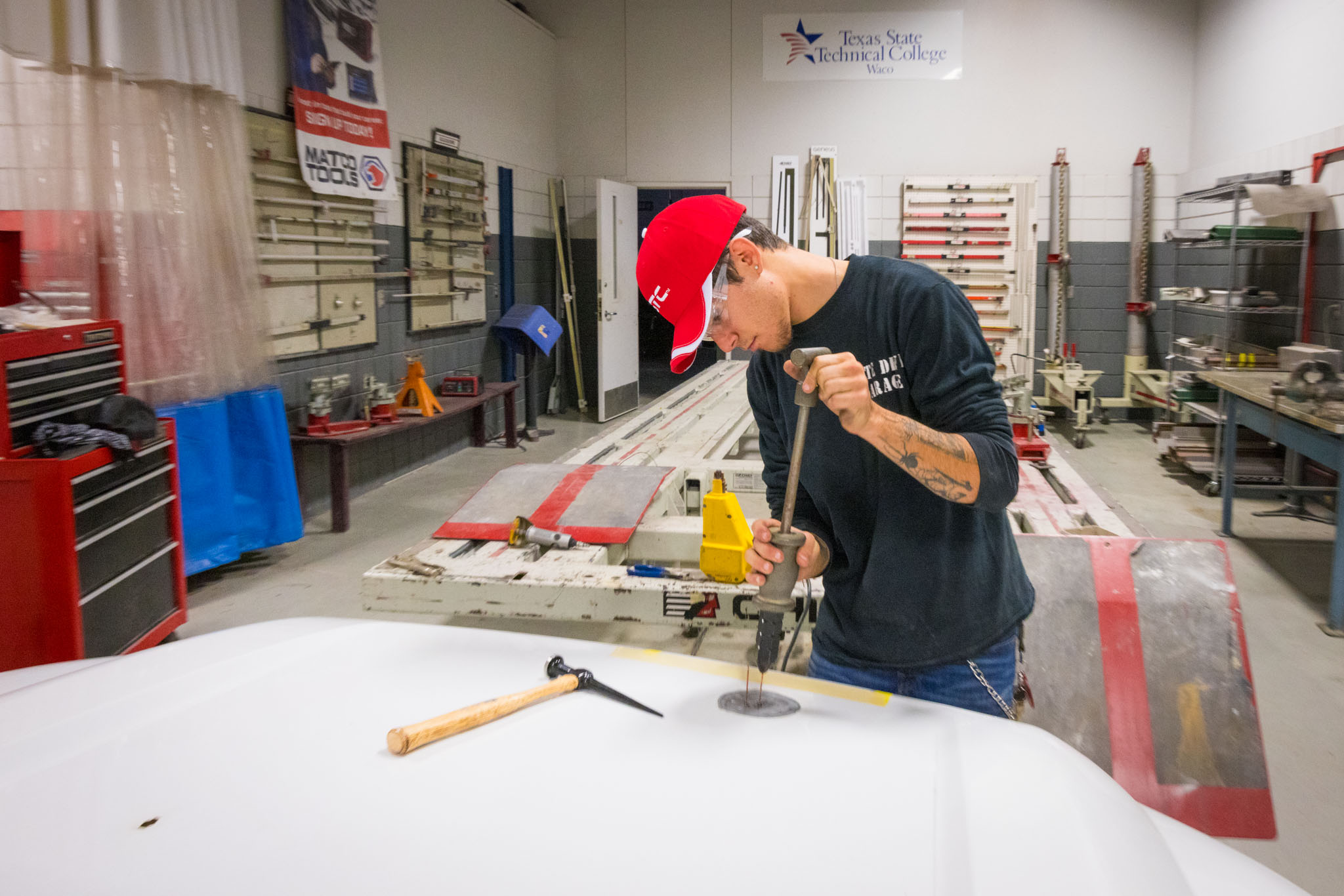 Student wearing a red cap uses a manual dent puller on a car panel, applying force to repair damage in an auto body training shop with tools and equipment in the background.
