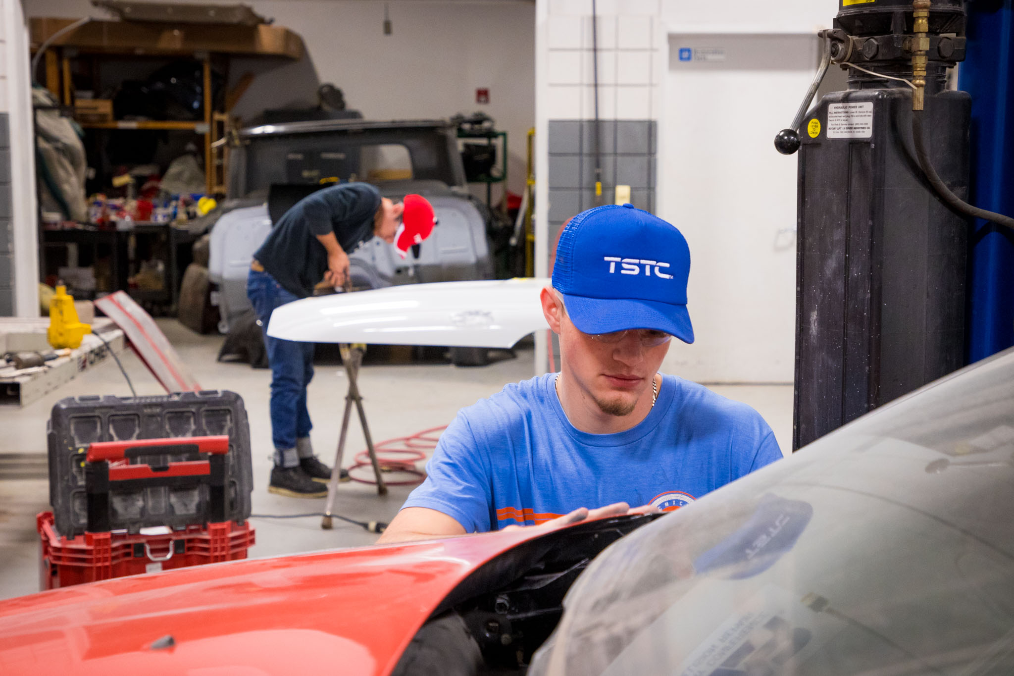 In an auto body lab, a student in a blue TSTC hat carefully adjusts a red car panel as another student works on a panel set on a stand in the background.