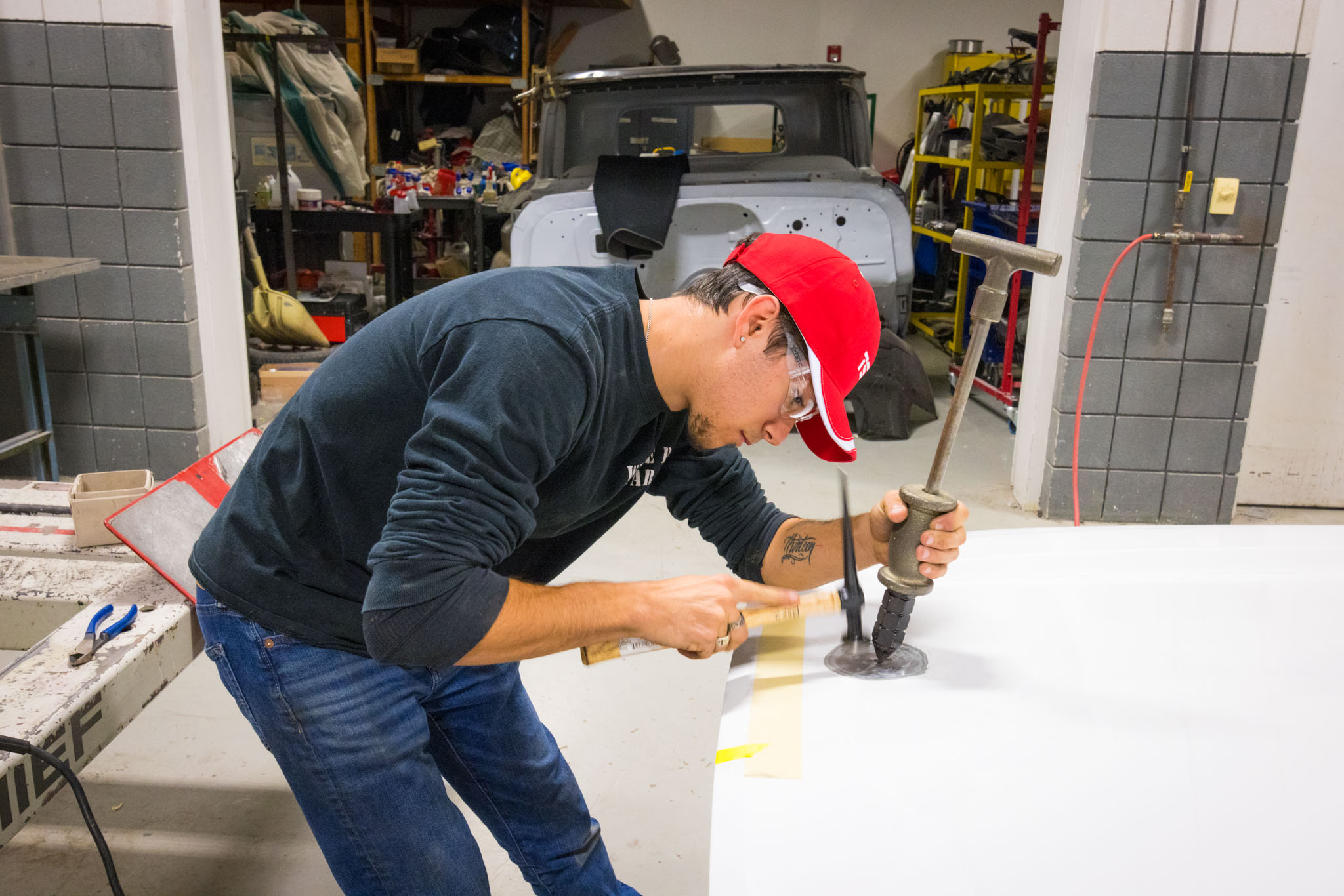 Inside an automotive lab, a student in a red cap carefully works a dent out of a car panel using a hammer and puller tool, with a partially restored vehicle behind him.