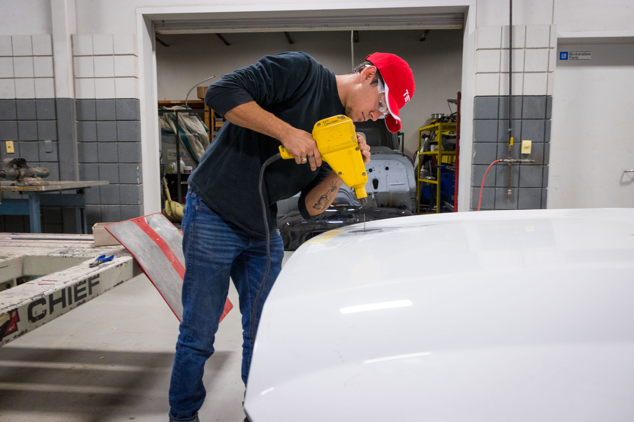 Inside an automotive lab, a student wearing a red cap carefully applies heat to a car panel with a handheld tool, with a partially restored vehicle visible behind him.