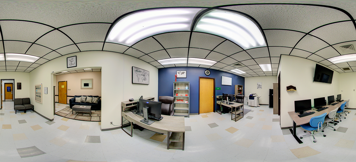 Panoramic view of an office-like workspace with desks and computers, a small seating area with a couch visible through an open doorway, shelving units, and a row of desktop stations along the wall under fluorescent ceiling lights.