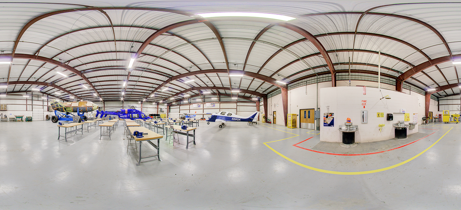 Panoramic view of an aviation training lab area with small aircraft and a helicopter, surrounded by worktables, tools, and open floor space inside a large industrial hangar.