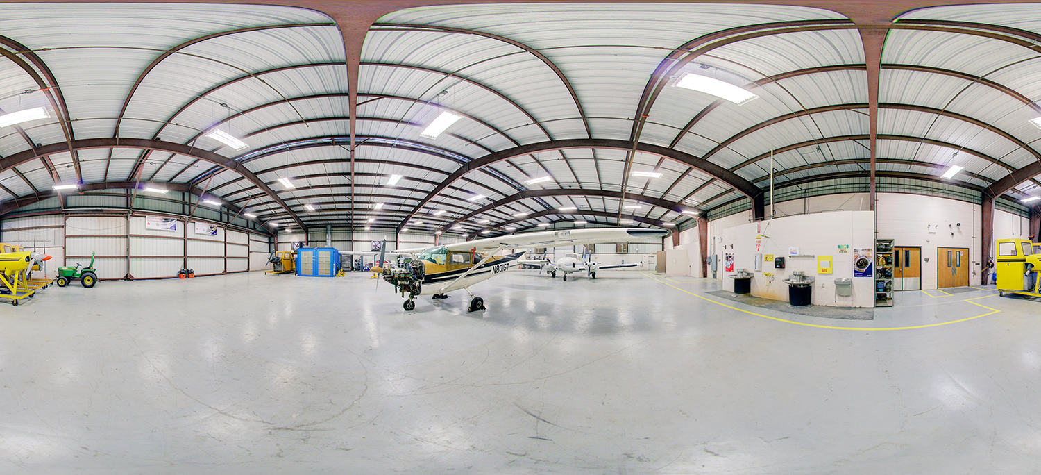 Panoramic view of an aviation hangar area with small airplanes parked on a wide open floor, surrounded by maintenance equipment, storage units, and workshop rooms along the walls.