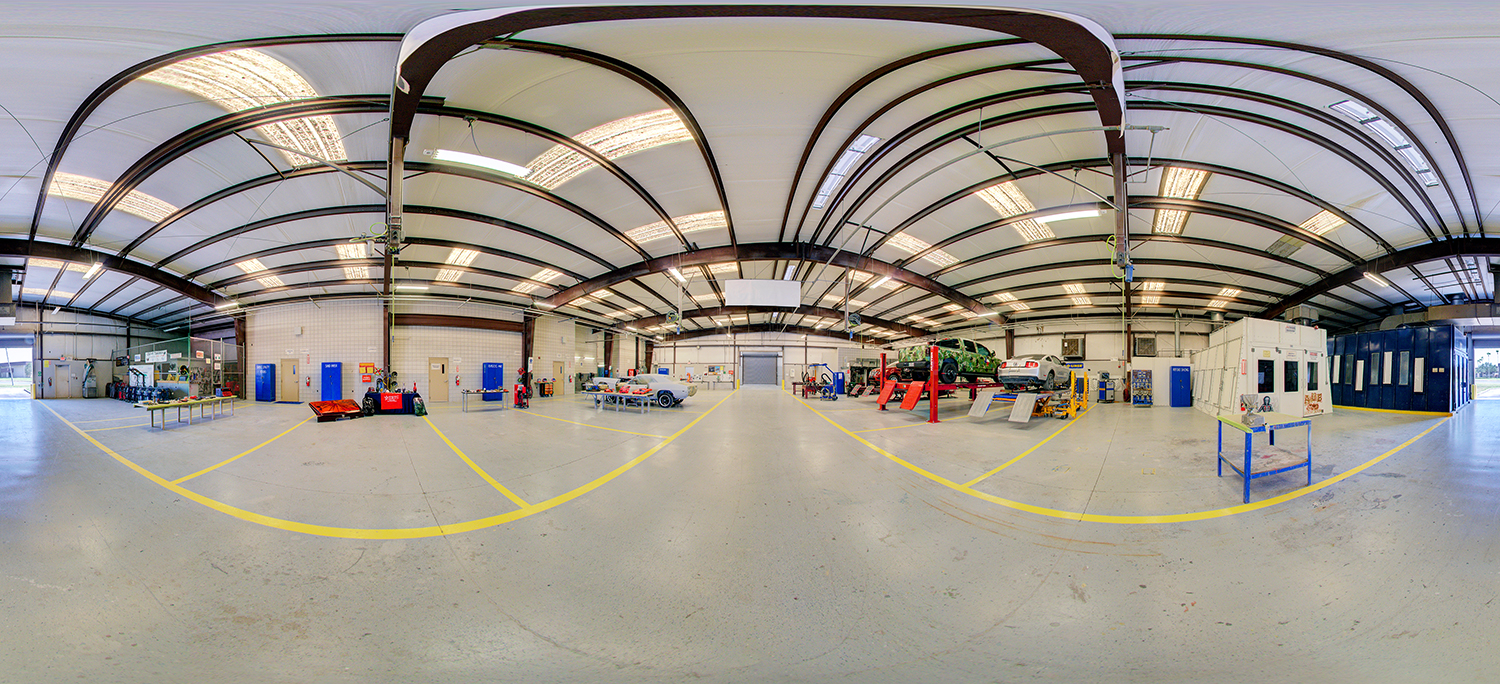 View of an autobody repair lab area with multiple vehicles on lifts, workstations, paint booths, and open floor space marked with yellow lines inside a large industrial training facility.