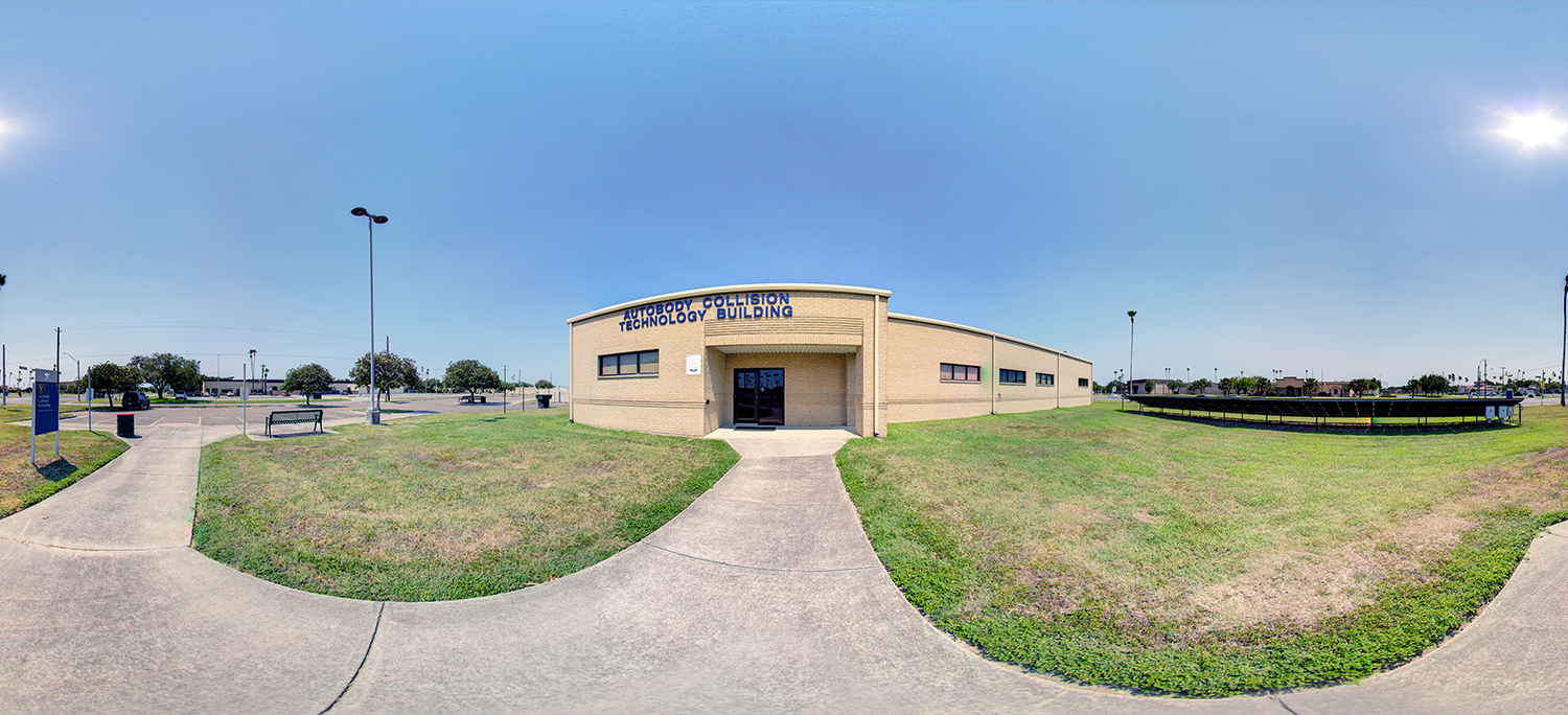 Panoramic view of an autobody collision technology building exterior with a curved brick facade, central entrance, surrounding grass lawn, sidewalks, benches, and a clear sunny sky.