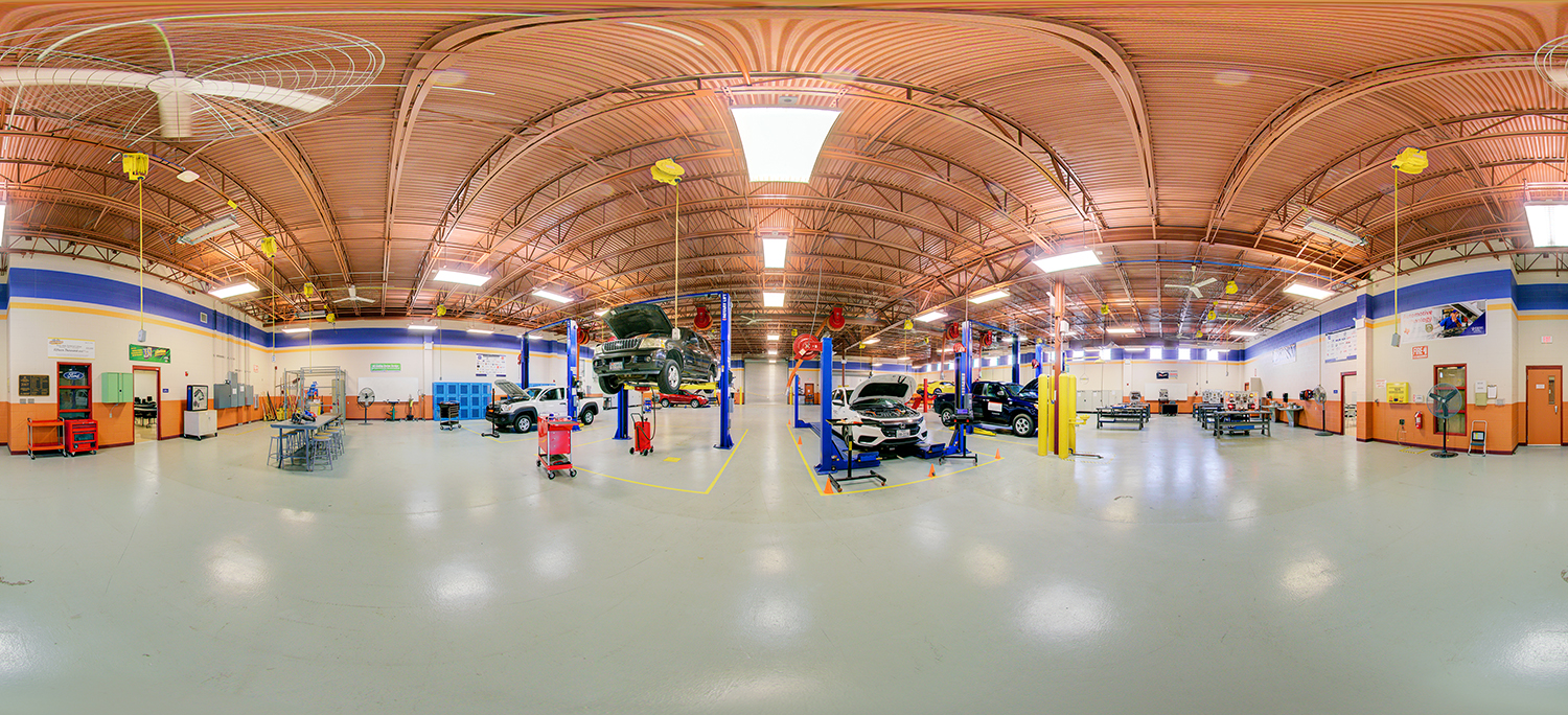 Panoramic view of an automotive training workshop with multiple cars on hydraulic lifts, tool stations, and work areas spread across a large, open garage space with high ceilings and bright industrial lighting.