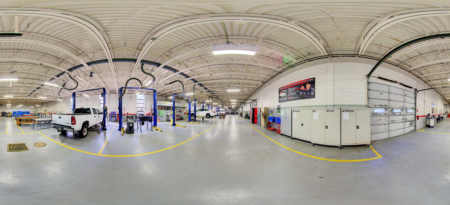 Panoramic view of a large automotive training lab with multiple vehicles on lifts, wide service bays marked by yellow lines, HVAC training units along one wall, and a roll-up garage door, all under a high industrial ceiling with exposed piping and bright overhead lights.