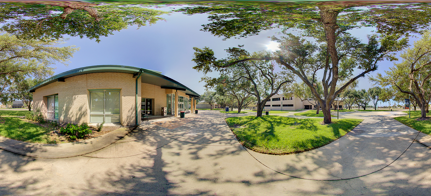 Panoramic view of an outdoor campus area with a small brick building, shaded walkways, trees casting shadows, green grass, and nearby academic buildings connected by paved paths.