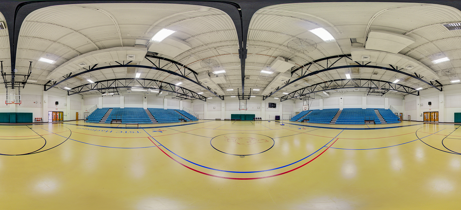Panoramic view of an indoor gymnasium with a polished court marked for basketball, high ceilings with exposed beams, multiple hoops, and rows of blue bleachers along both sides.