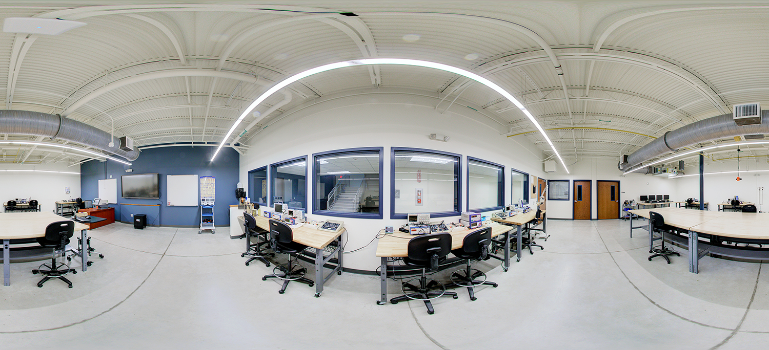 Panoramic view of an electronics lab area with workbenches, computers, and testing equipment arranged around the room, featuring interior observation windows, ceiling-mounted lighting and ductwork, and adjacent classrooms.