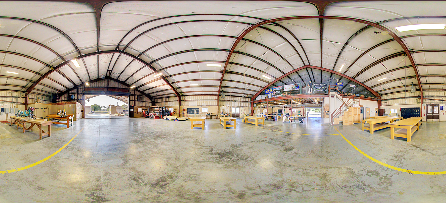 Panoramic view of a large workshop area with multiple wooden workbenches, tools, and equipment, featuring a high metal ceiling, an open garage door, and a mezzanine level with stairs and storage space.