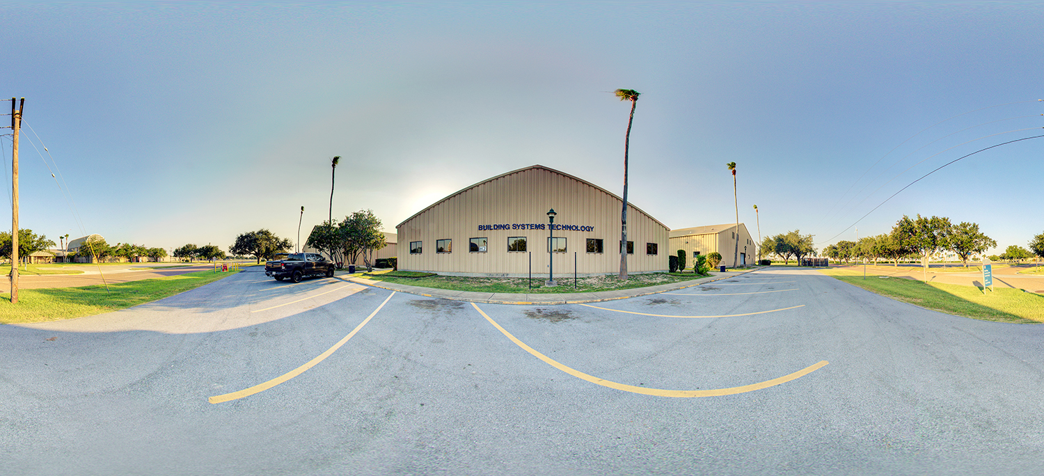 Panoramic view of a building labeled “Building Systems Technology,” with a parking area in front, a few parked vehicles, tall palm trees, and surrounding grassy campus area under a clear sky.