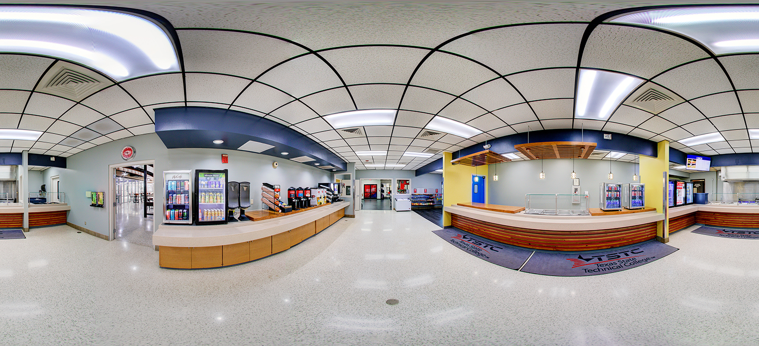 Panoramic view of a campus cafeteria or food service area with long service counters, beverage coolers, coffee machines, and a serving station beneath pendant lights, set in a bright, open space with tiled flooring and overhead fluorescent lighting.
