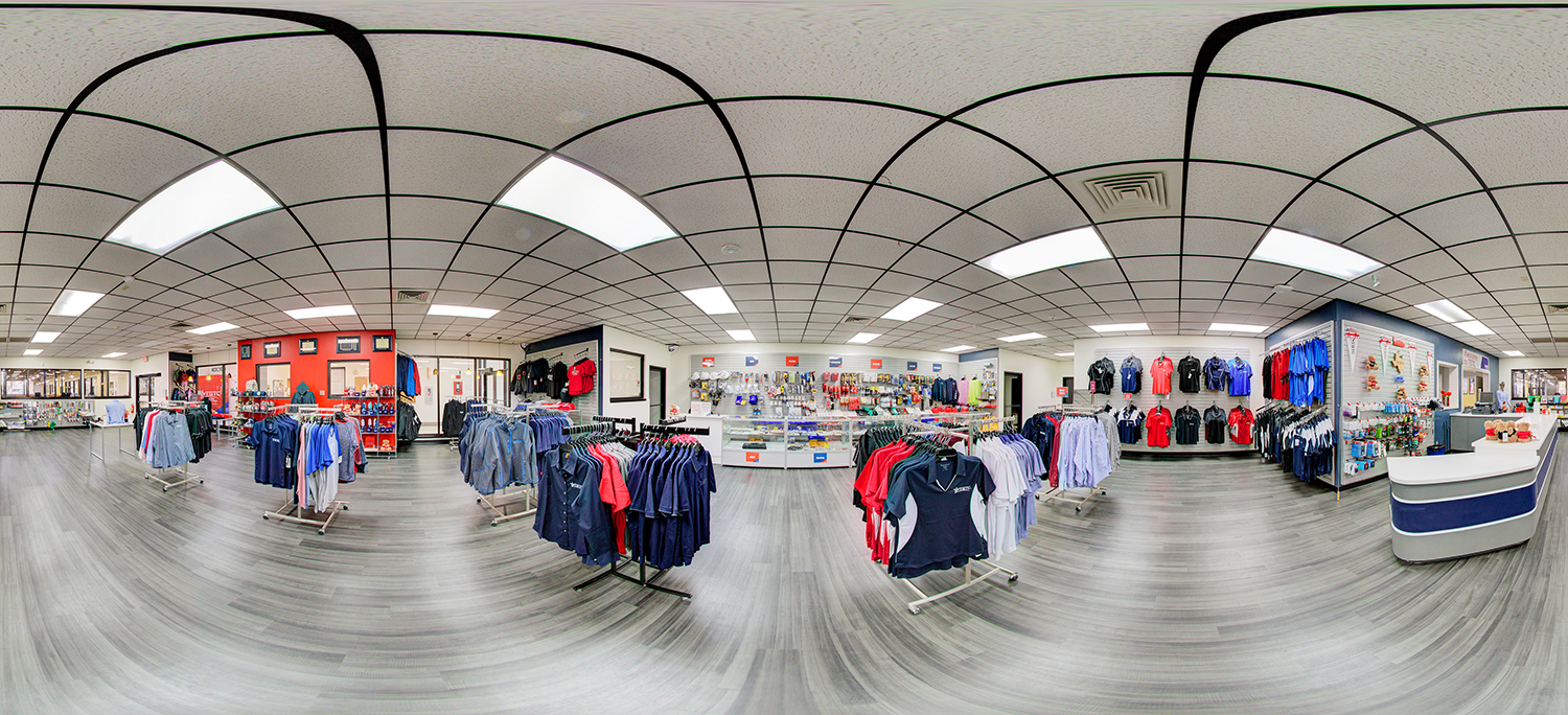Panoramic view of a campus retail store featuring racks of clothing in red, blue, and neutral tones, wall displays of apparel and accessories, glass display cases, and a checkout counter on a wood-look floor under grid ceiling lights.