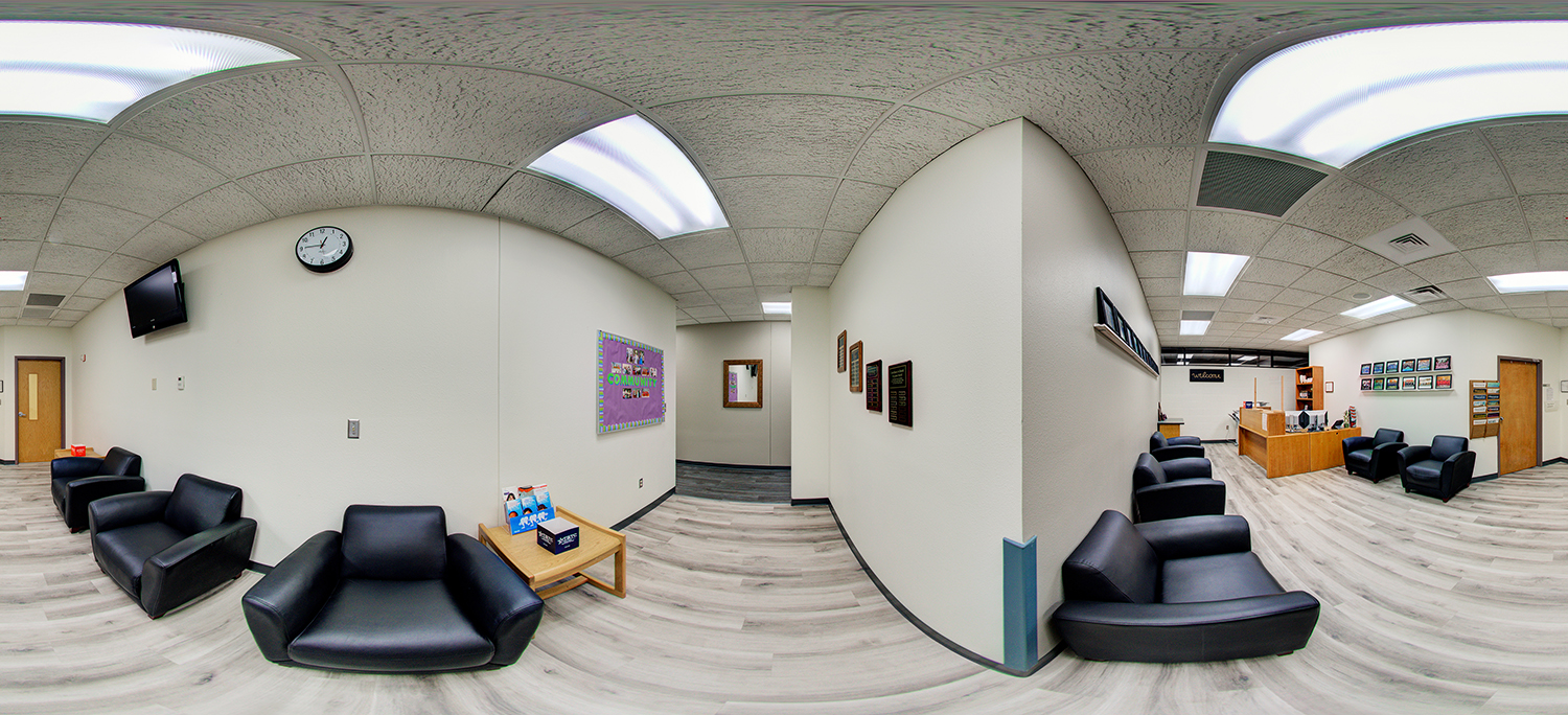 Panoramic view of an indoor reception and waiting area with black lounge chairs, a small table with brochures, a front desk, wall plaques, and a hallway leading to offices under bright fluorescent lighting.