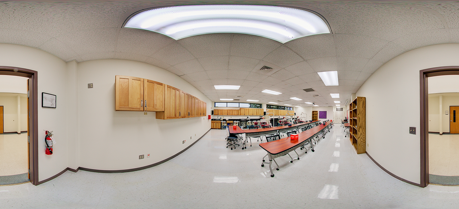 Panoramic view of a clinical training classroom with long curved tables, rolling chairs, and rows of medical practice stations at the back, along with wooden cabinets, shelving, and bright overhead lighting.