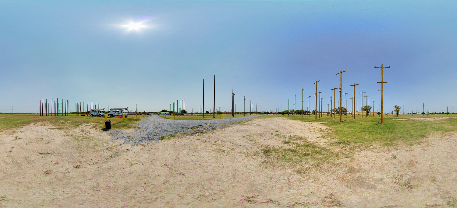Panoramic view of an outdoor utility training area with numerous wooden utility poles arranged across a wide open field, gravel paths, work vehicles, and clear sky overhead.