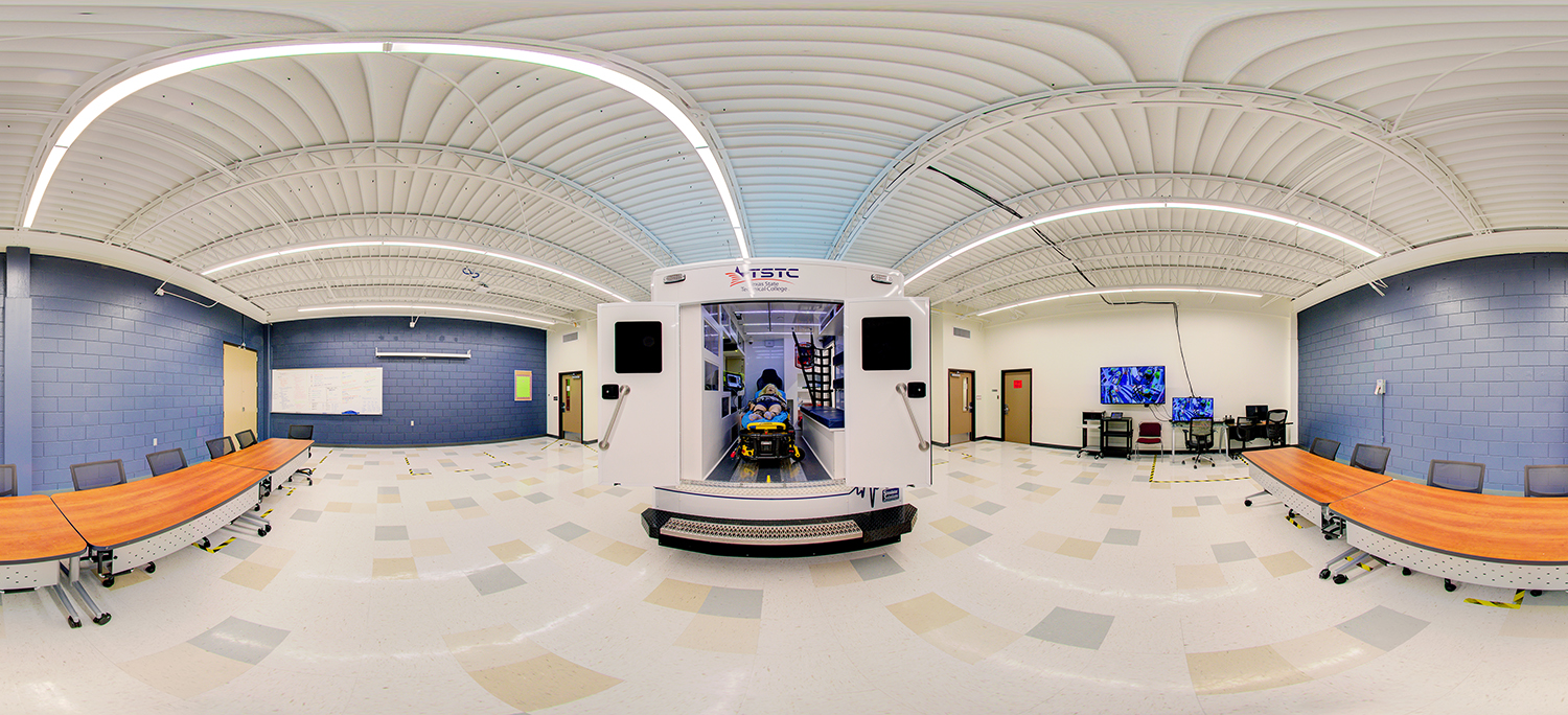 Panoramic view of a medical training classroom featuring a central ambulance simulator with a stretcher and mannequin inside, surrounded by tables, chairs, whiteboards, and computer stations along blue and white walls.