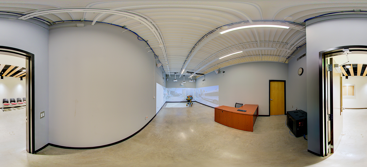 Panoramic view of a simulation lab with a stretcher and medical mannequin centered in front of large projection screens, a small instructor desk to the side, and adjacent rooms visible through open doorways.
