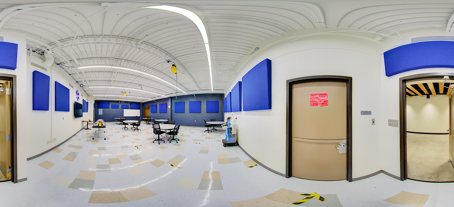 Panoramic view of a medical training classroom with blue acoustic wall panels, round tables and chairs, a mannequin on a stretcher, and a supply cart, with doors leading to adjacent rooms and marked floor spacing guides.