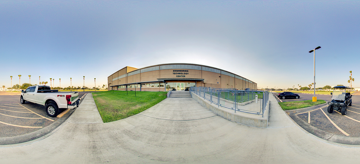 Panoramic view of a building labeled “Engineering Technology Center,” with a concrete walkway and accessibility ramp leading to the entrance, surrounded by parking areas, vehicles, and open grassy campus grounds.