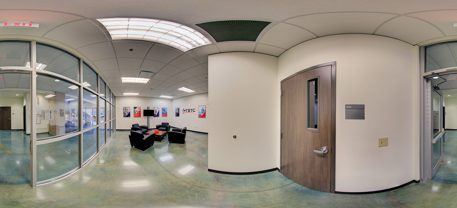 Panoramic view of an indoor hallway lounge area with glass-walled offices on one side, a cluster of black armchairs around a small table, wall posters, and a door labeled “101K,” all under bright ceiling lights.