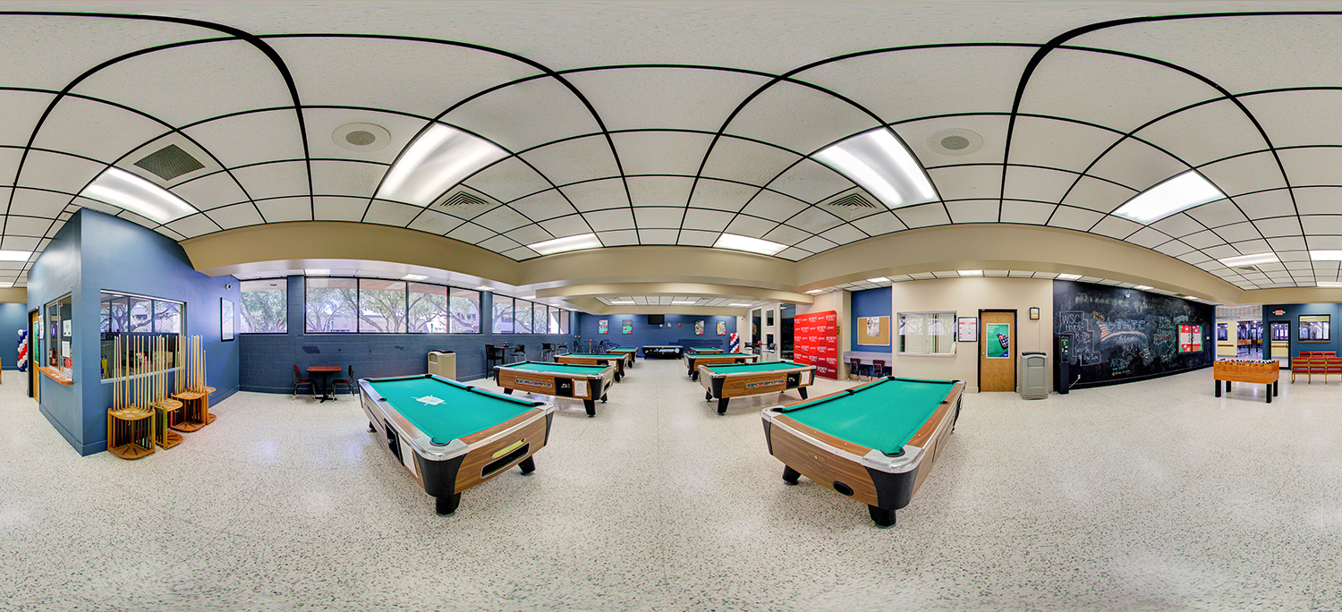 Panoramic view of a student recreation or lounge area featuring multiple pool tables arranged across the room, cue racks along the wall, a foosball table, and seating areas, with large windows and a chalkboard wall under bright ceiling lights.