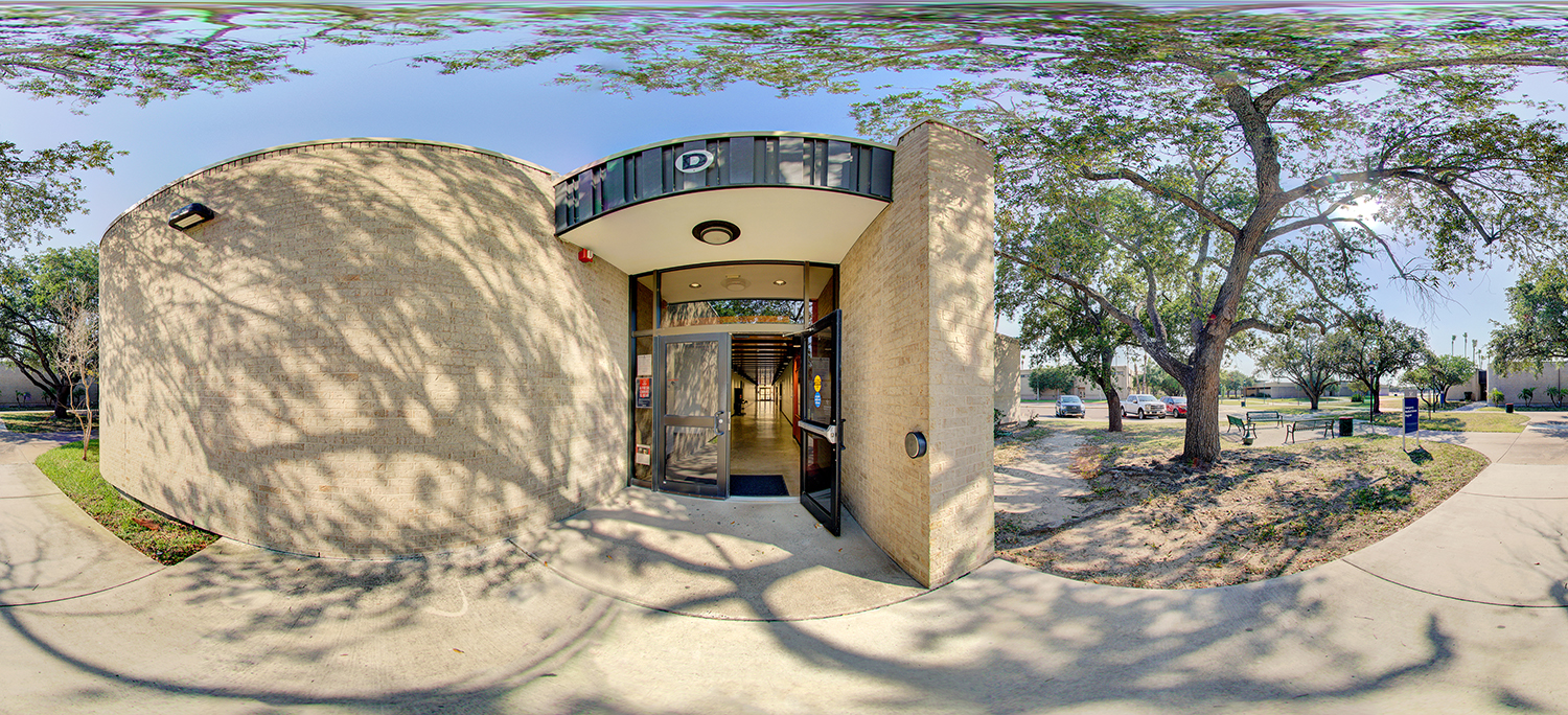 Panoramic view of a curved building entrance with glass doors propped open, leading into a hallway, surrounded by trees casting shadows, nearby benches, and campus walkways under a clear sky.