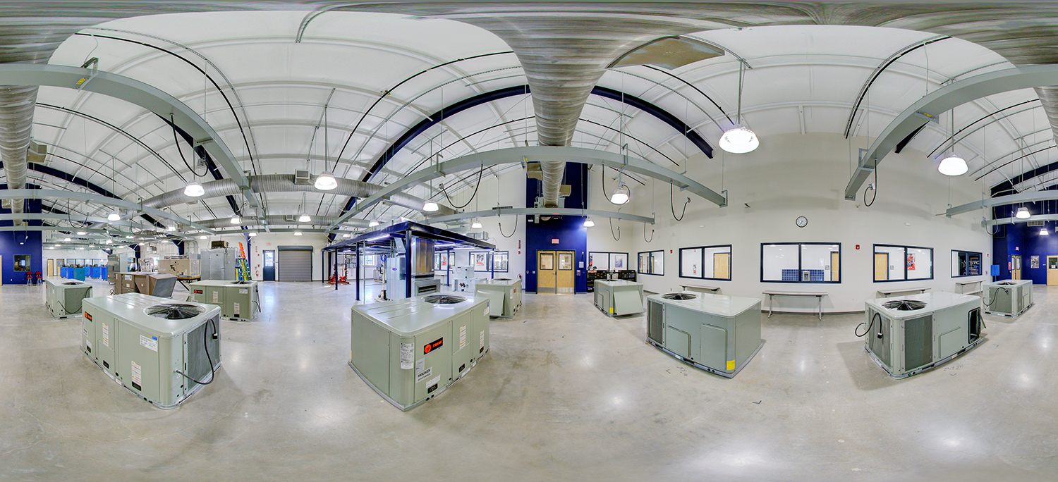 Panoramic view of an HVAC training lab area with multiple large air conditioning units arranged across the floor, exposed ceiling ductwork and piping, and classroom windows and doors along the walls.